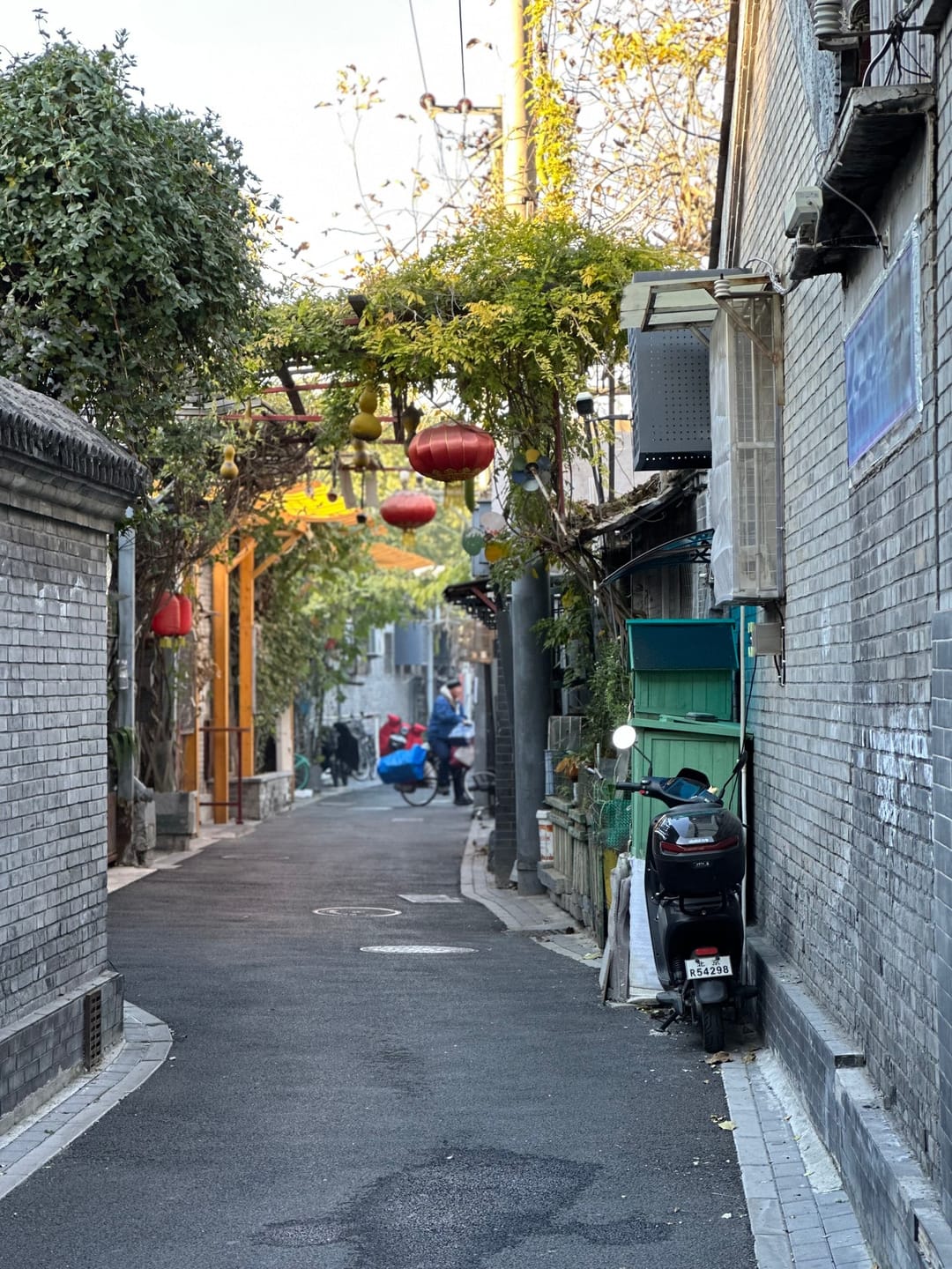 A narrow hutong lane in Dongsi with grey brick walls, red lanterns, climbing plants and scooters parked along the side