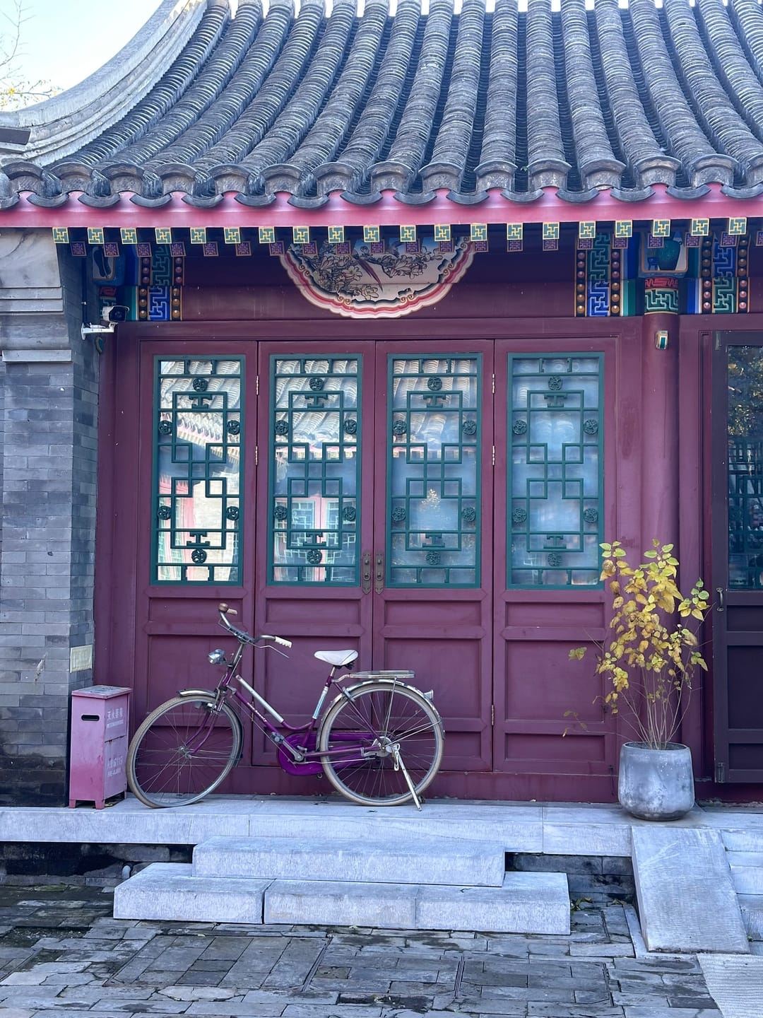 Ein rotes Holztor mit Gitterfenstern in einem Hutong, davor ein abgestelltes Fahrrad und Topfpflanzen