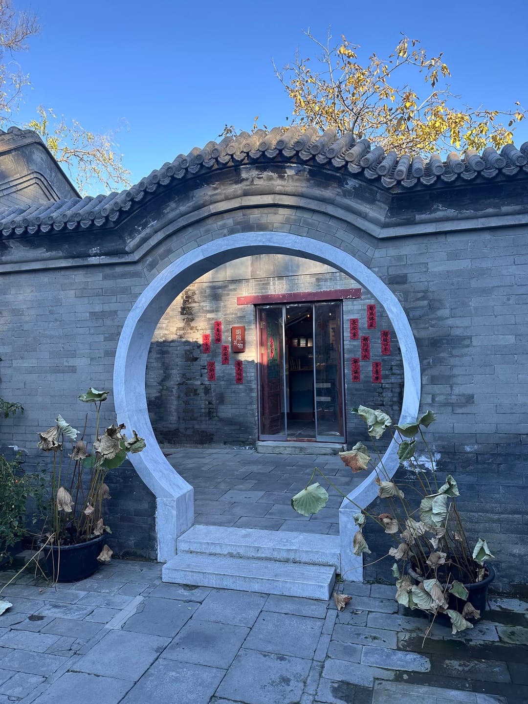 Un portale rotondo in un muro grigio di un hutong, oltre il quale si vede un cortile con una porta rossa