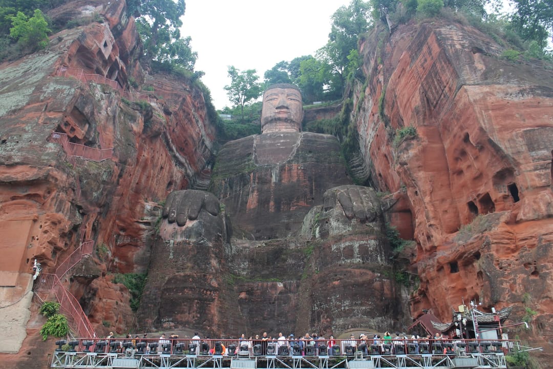El Buda Gigante de Leshan sentado en un acantilado de arenisca roja, con árboles en la cabeza y gente a sus pies