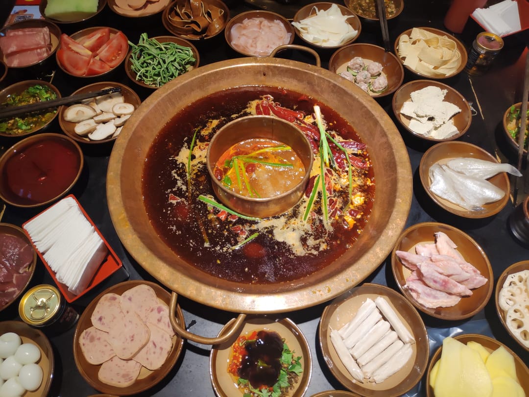 Olla de cobre con un anillo de caldo rojo de chile alrededor de un centro más suave, entre platillos de carne, tofu y verdura