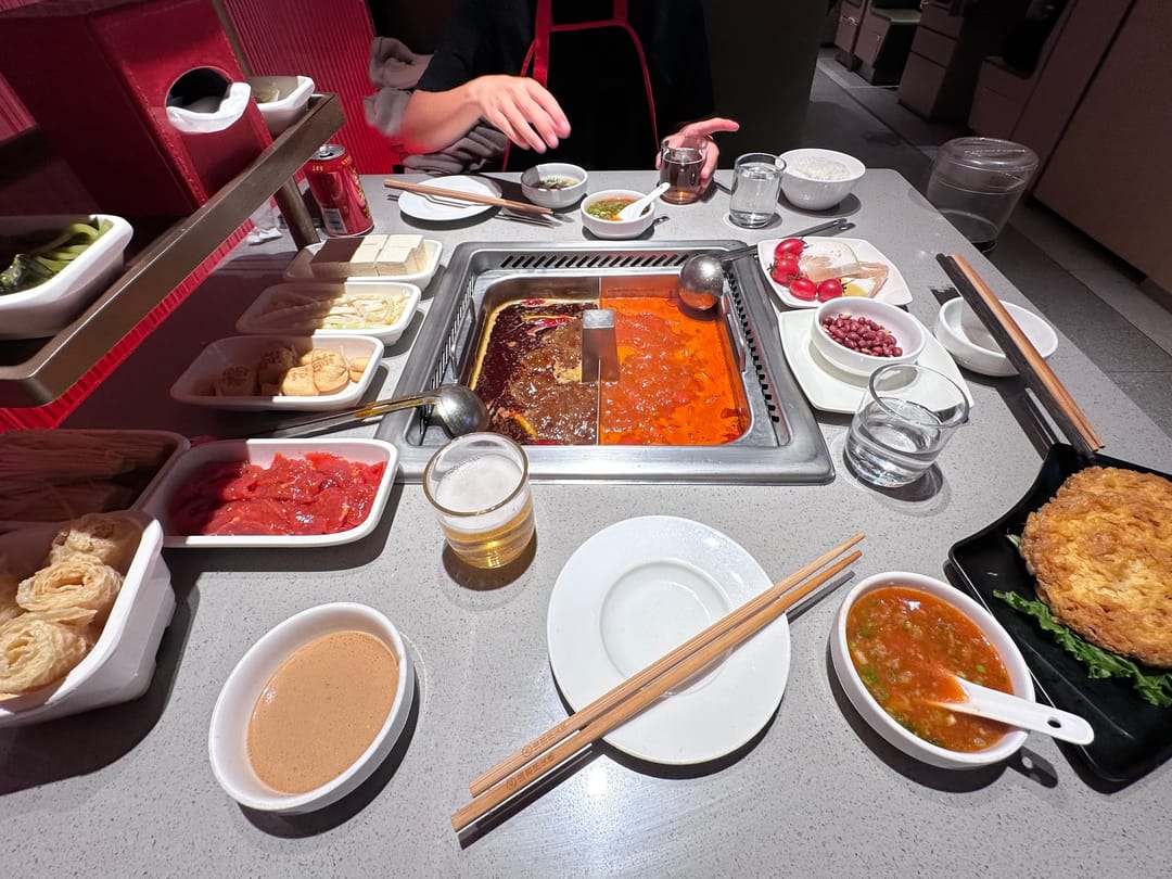A hotpot table in Beijing with a divided pot of red spicy and clear broth, plates of raw meat and vegetables, dipping sauces and a glass of beer