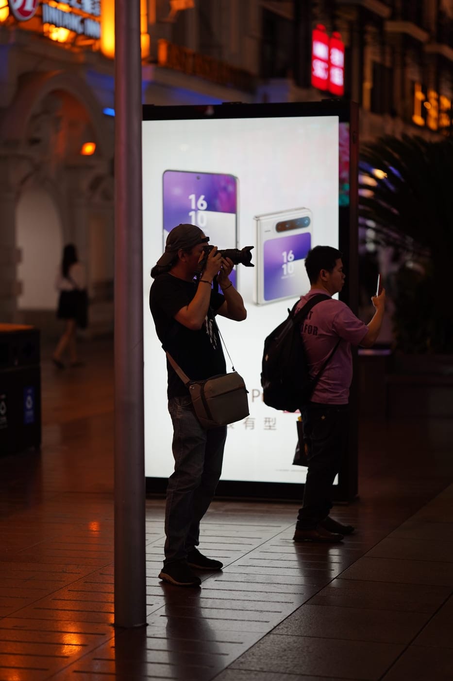 Two people photographing at night on Nanjing Road in Shanghai, in front of a lit smartphone advertisement