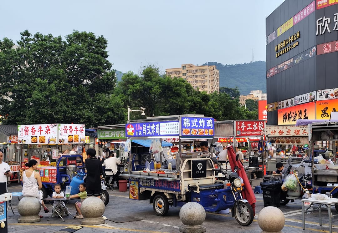 Banchi di cibo da strada su tricicli elettrici in una piazza di Shenzhen, con insegne colorate e clienti seduti ai tavolini
