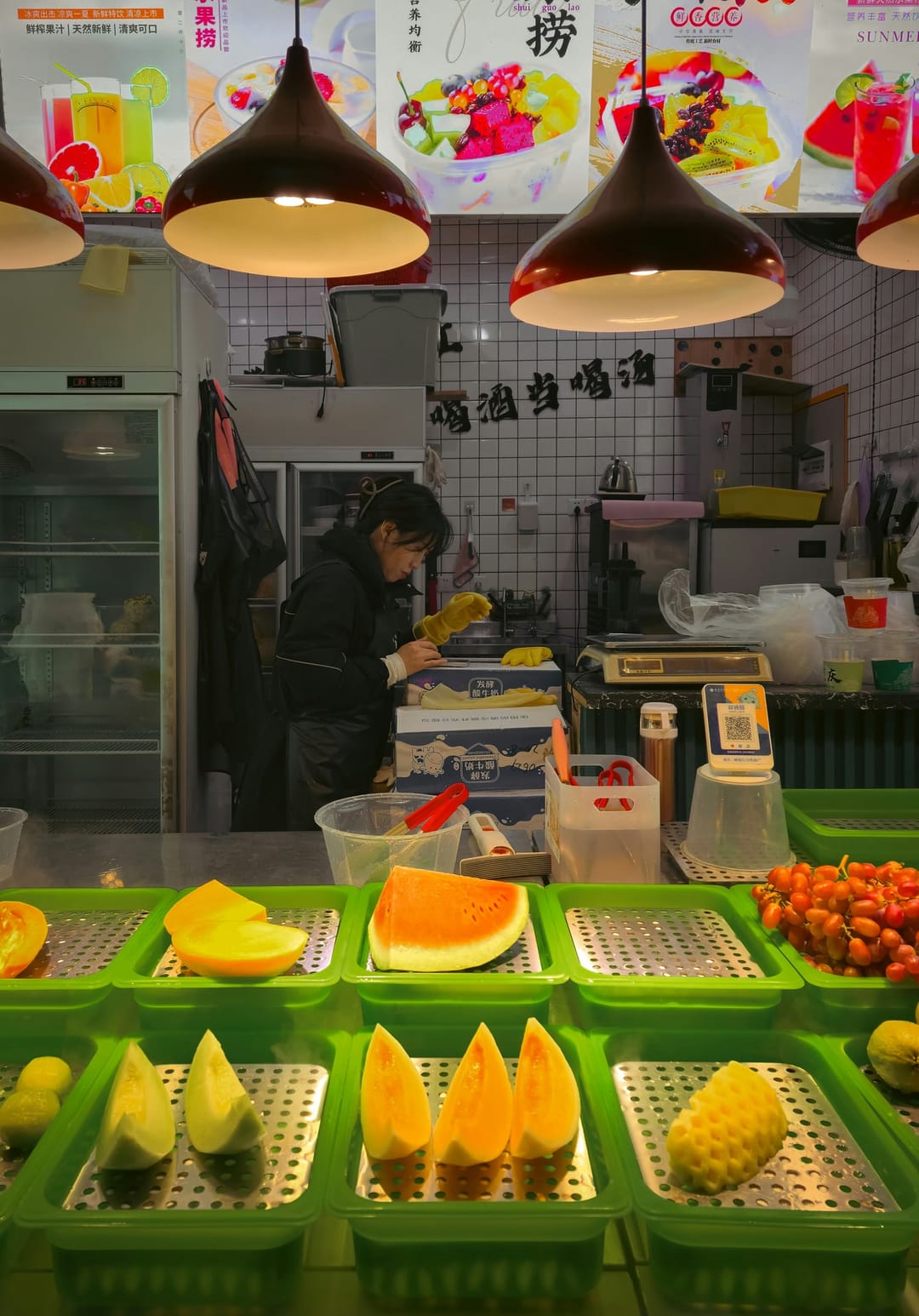 A fruit stall in Chongqing with cut melon and papaya in green trays, a scale and the vendor working behind the counter