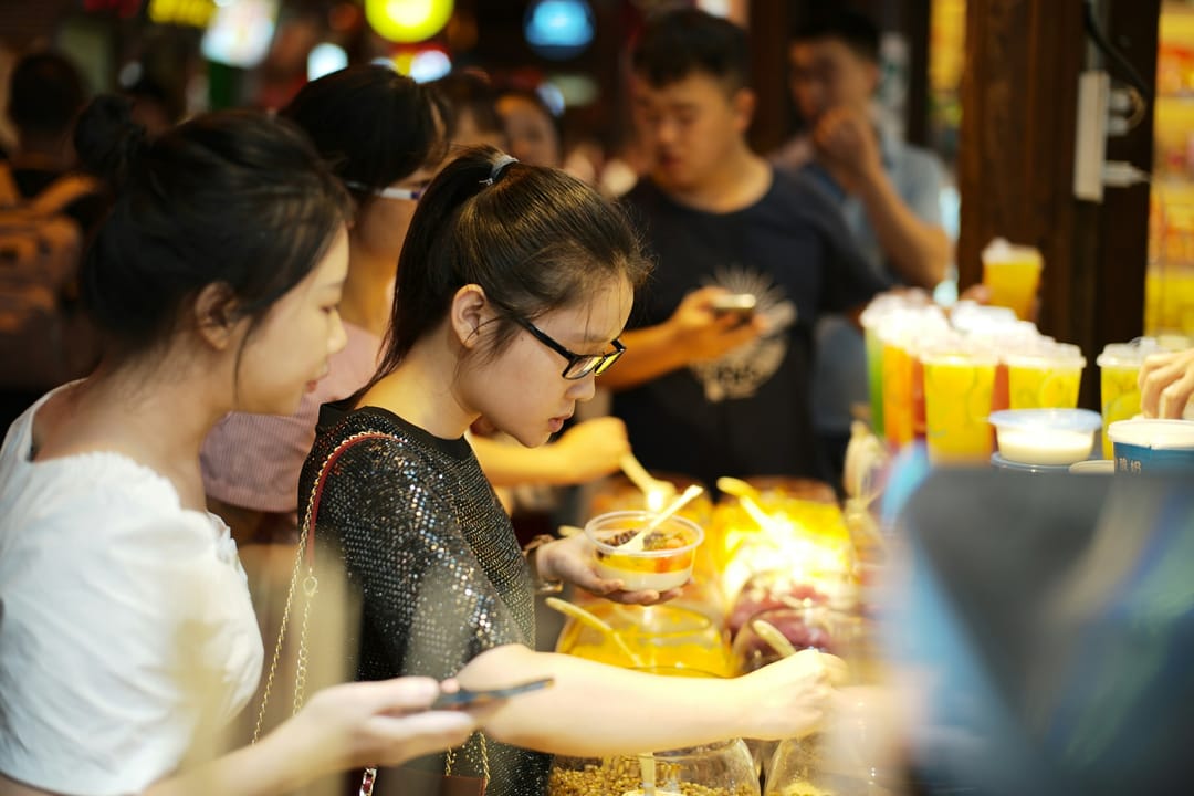 Customers at a night-market stall in Chongqing choosing desserts from trays, one of them holding a phone