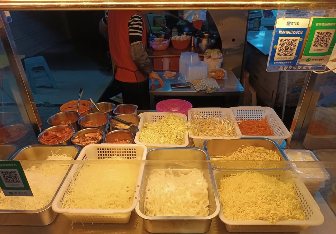 Nudelstand auf einem Nachtmarkt in Shenzhen mit Körben voller Nudeln und Gemüse, oben rechts die Zahlungscodes von Alipay und WeChat Pay