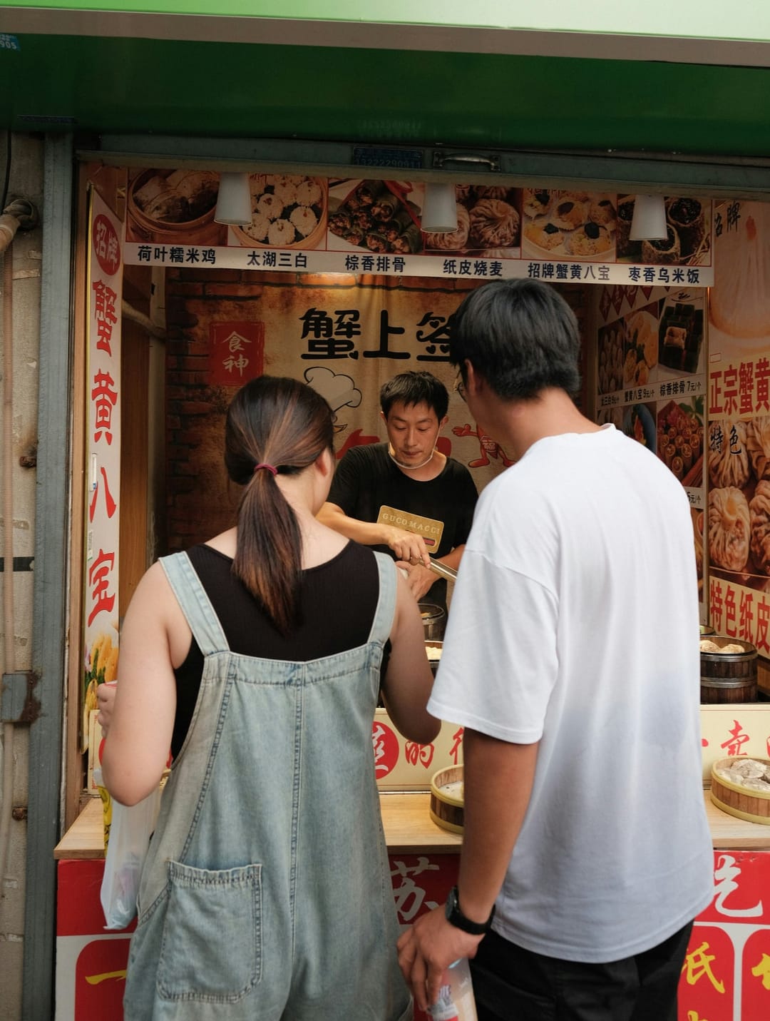 Two customers in front of a dumpling counter in Suzhou while the vendor serves them, with menu boards above