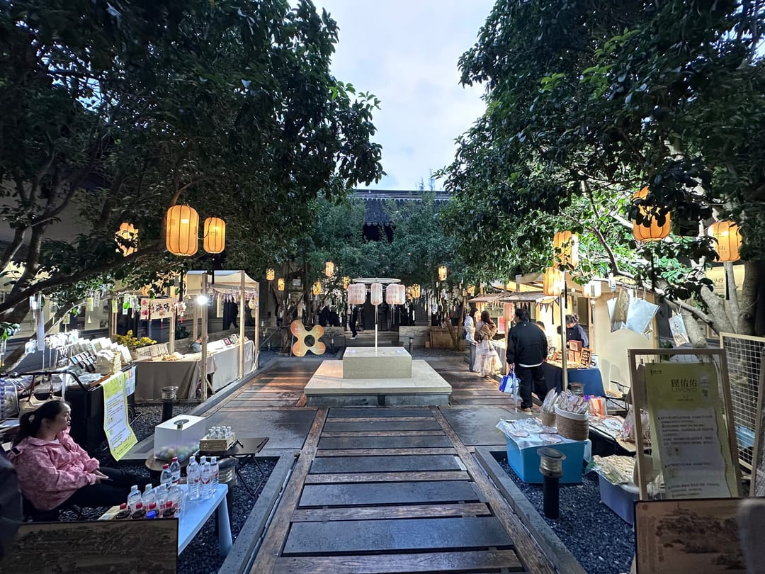 Bancarelle di artigianato sotto lanterne accese in un cortile alberato di Shantang Jie a Suzhou, al tramonto, con alcuni passanti