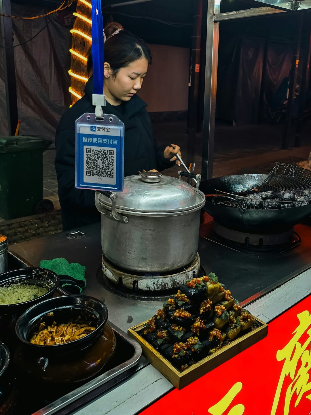 A cook at a street-food stall in Zhangjiajie, with an Alipay QR code hanging from a blue lanyard above the pots