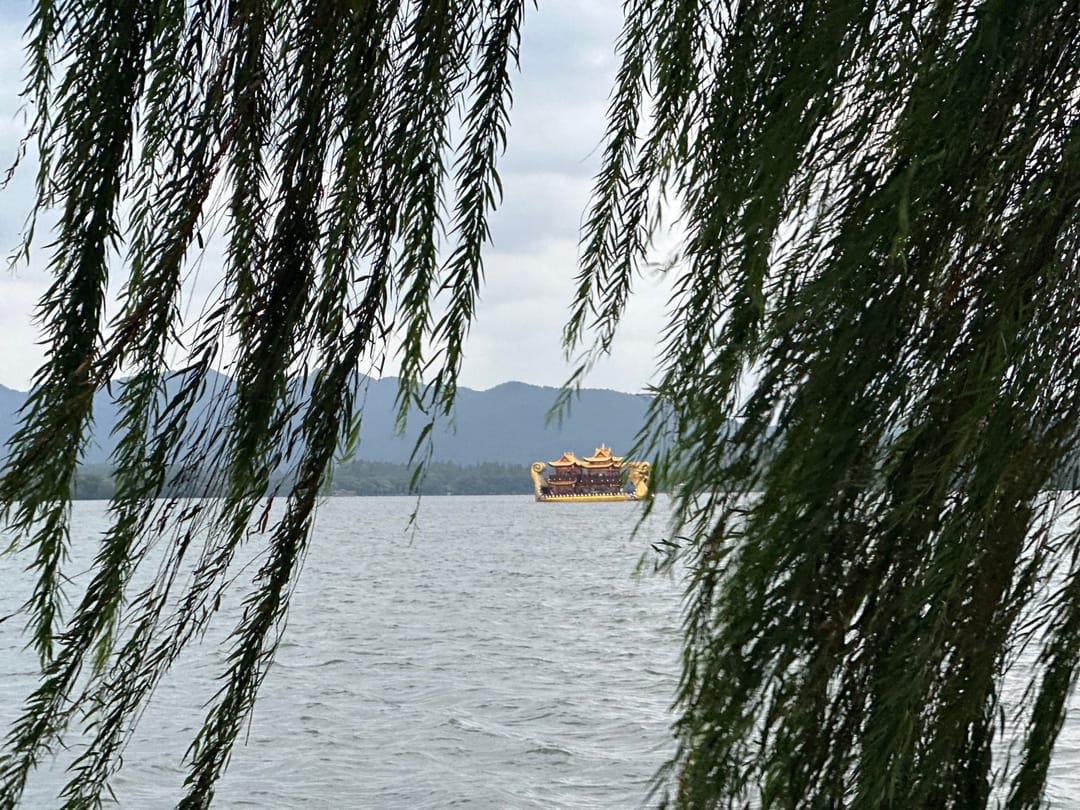 Un battello dorato in stile antico sul Lago dell’Ovest a Hangzhou, visto tra i rami di un salice piangente, con le colline sullo sfondo