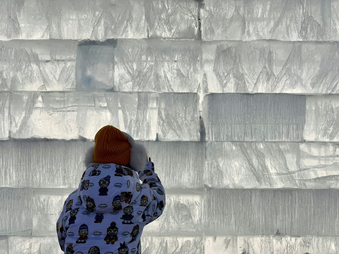 Ein Kind in Winterjacke und Mütze vor einer Mauer aus großen Eisblöcken in Harbin
