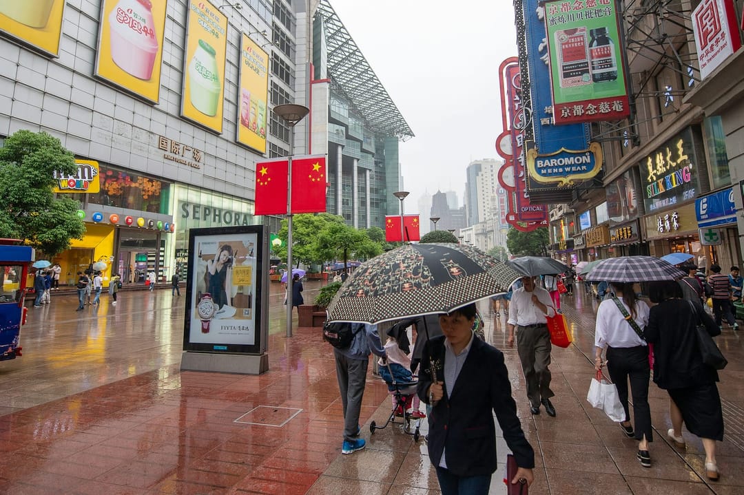 Shoppers with umbrellas walking along a wet pedestrian street in Shanghai lined with neon signs