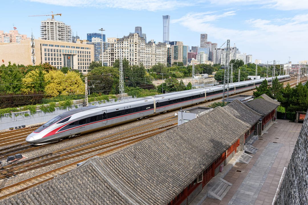 A white and red Fuxing high-speed train running past grey-tiled courtyard roofs in central Beijing, with office towers behind