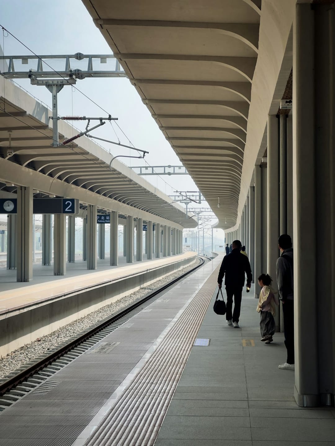 Ein Vater mit Tasche und ein kleines Kind gehen unter dem geschwungenen Dach den Bahnsteig 2 des Bahnhofs Jining Nord entlang
