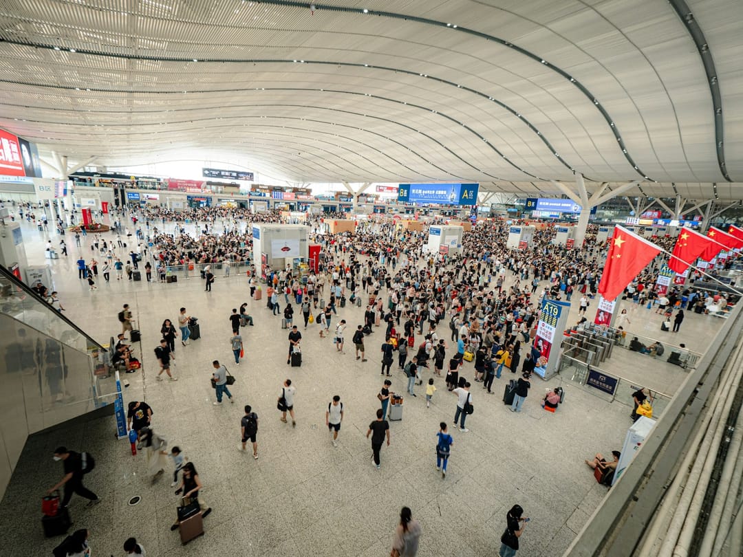 A crowded concourse at Shenzhen North railway station seen from above, with travellers pulling suitcases between the gates and red flags on the right