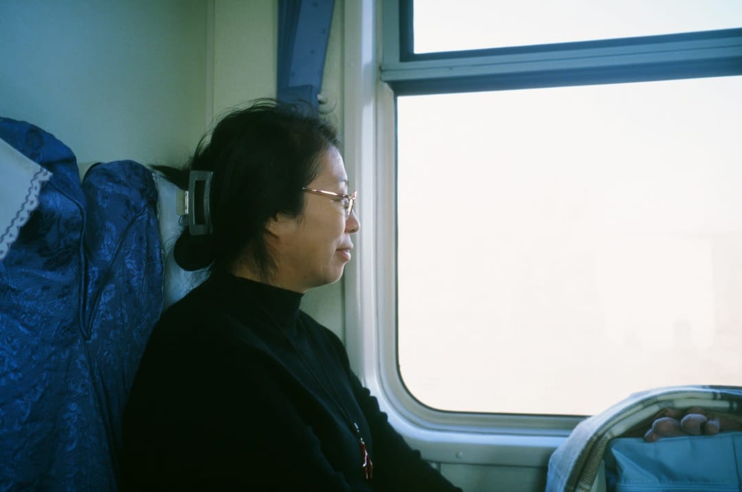 Eine Frau mit Brille sitzt am Fenster eines klassischen chinesischen Zugs mit blauen Sitzen und schaut hinaus