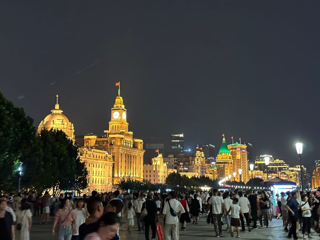 Crowds on the Bund promenade in Shanghai at night, the illuminated clock tower and domed buildings behind them