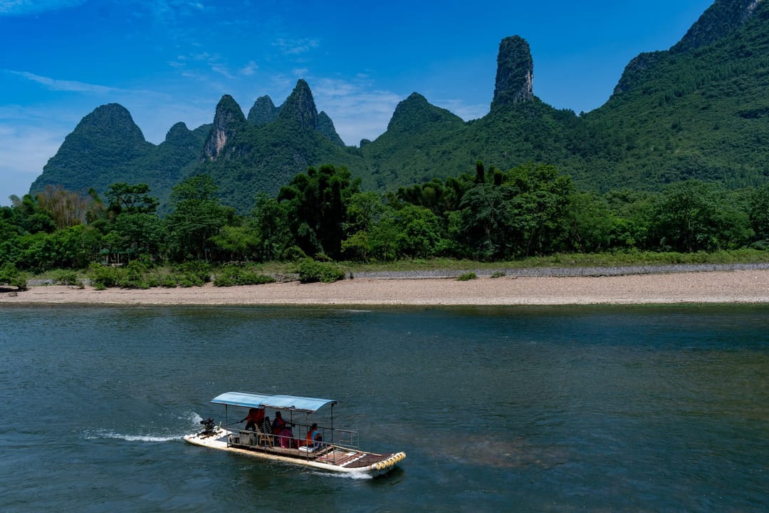 A bamboo raft carrying passengers on the Li River, green karst peaks and a pale sand bank behind it