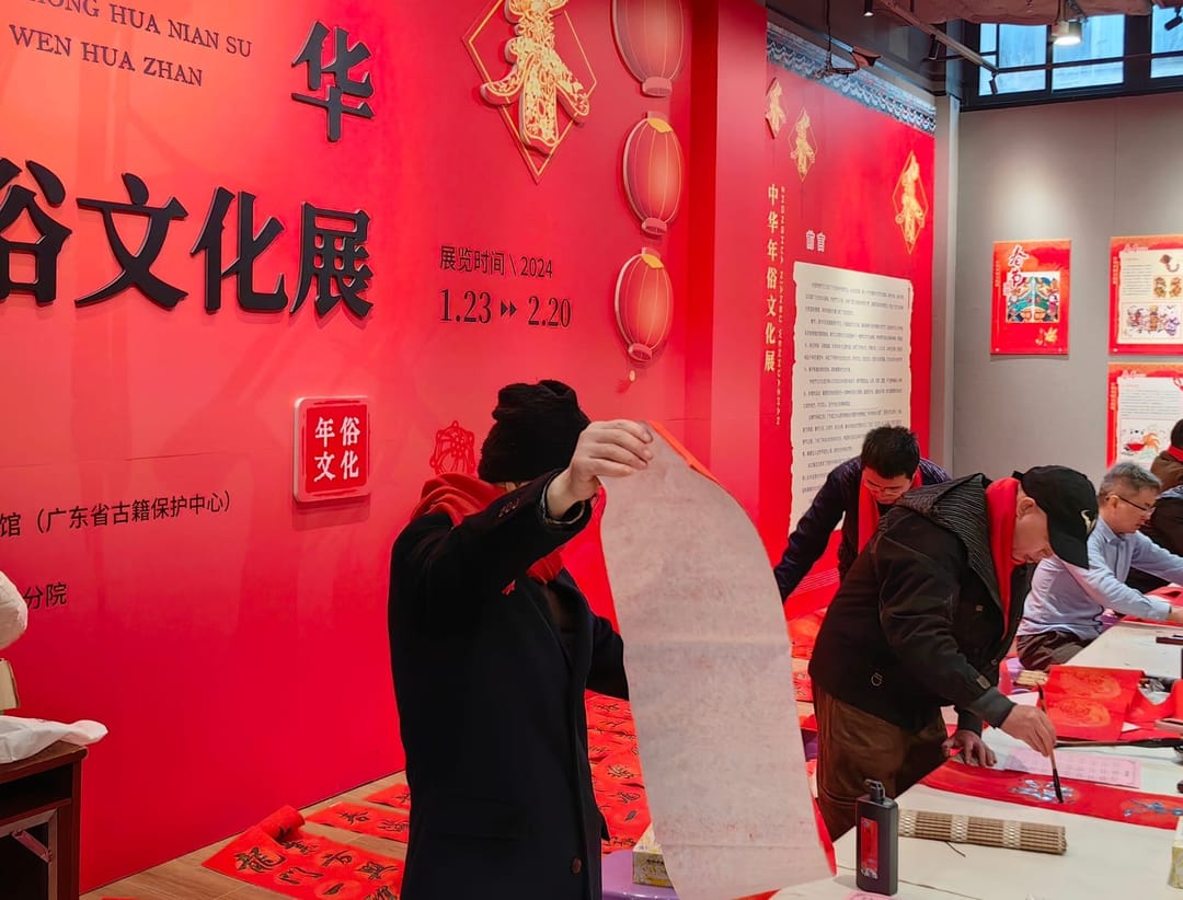 Volunteers writing red New Year couplets with brushes at long tables while a man holds up a long paper scroll