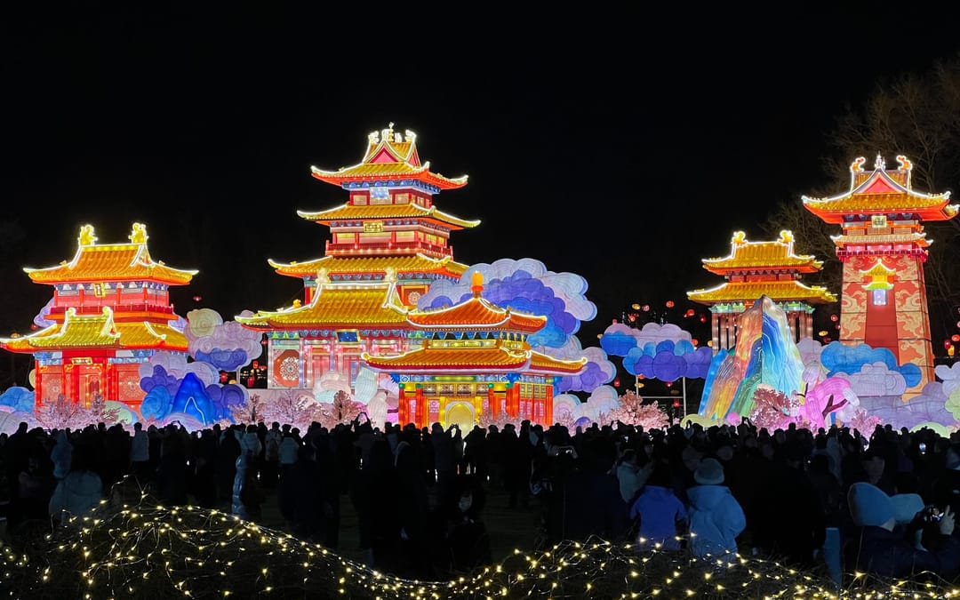 Lantern pavilions and painted clouds lit up at night at the Old Summer Palace, with a dark crowd watching in front