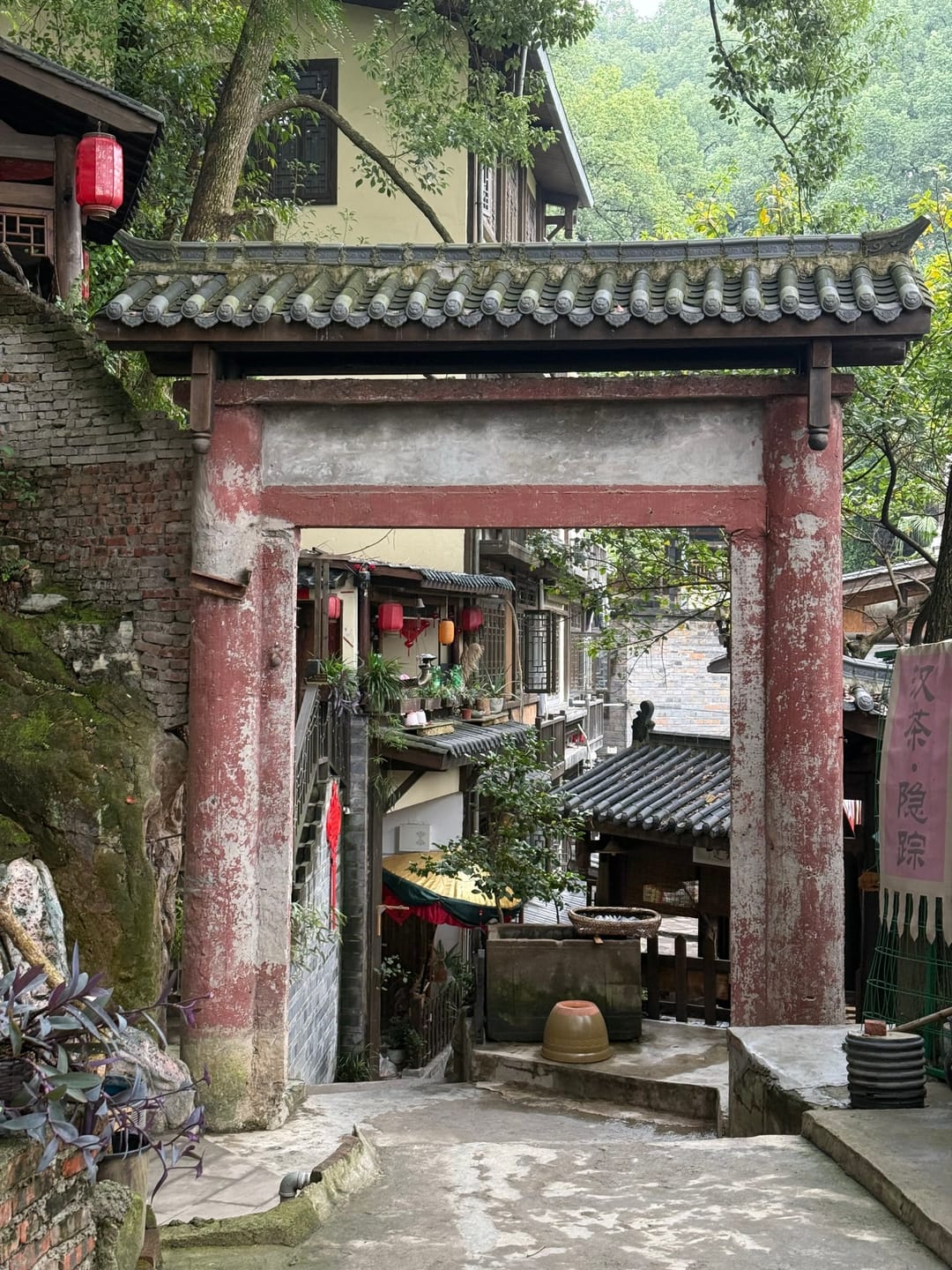 Un portal de piedra gastada con columnas rojas en el antiguo sendero de Huangge, con farolillos en las casas de la ladera