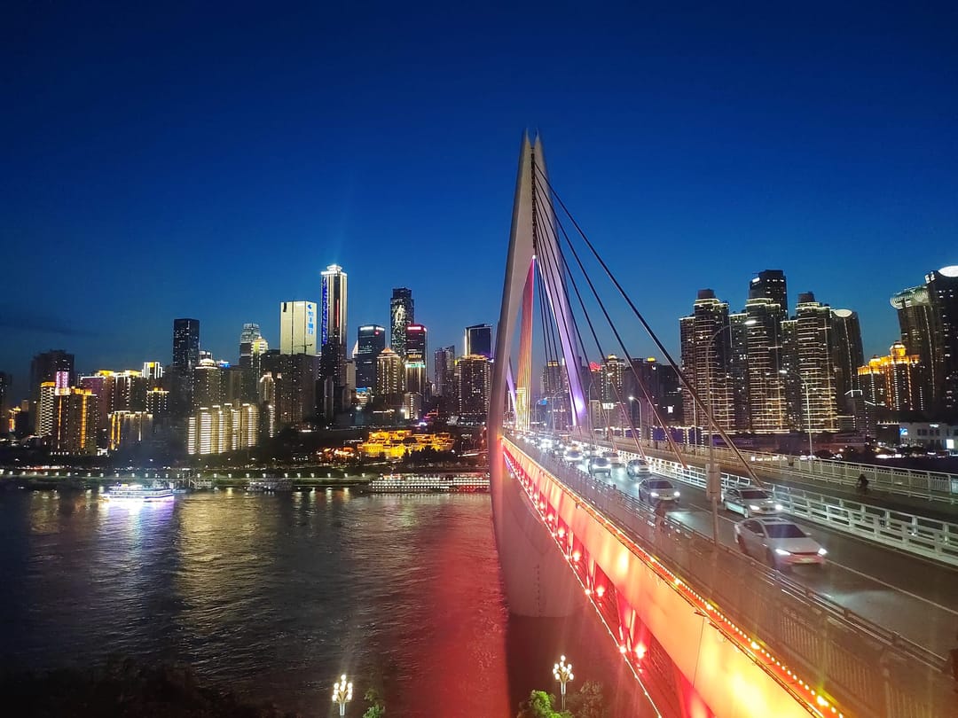 El tablero iluminado de rojo y el pilono blanco del puente de Qiansimen al anochecer, con las torres de Yuzhong al otro lado