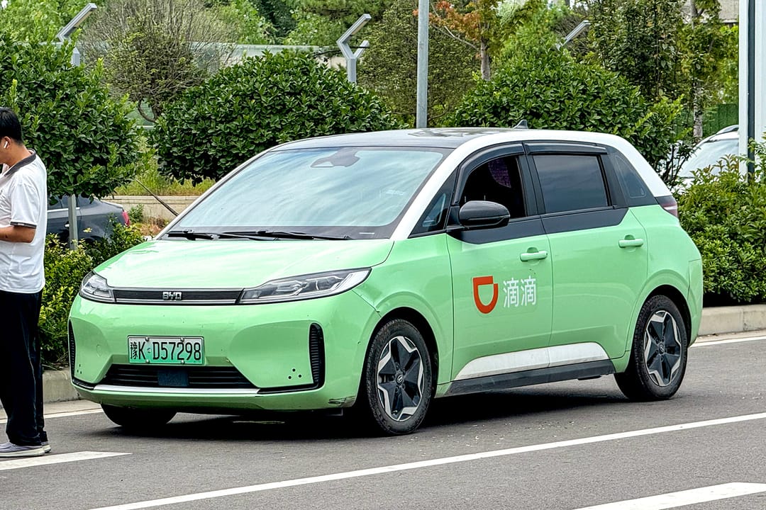 Un coche eléctrico BYD D1 verde claro con el logotipo naranja de DiDi en la puerta, aparcado ante una estación de tren