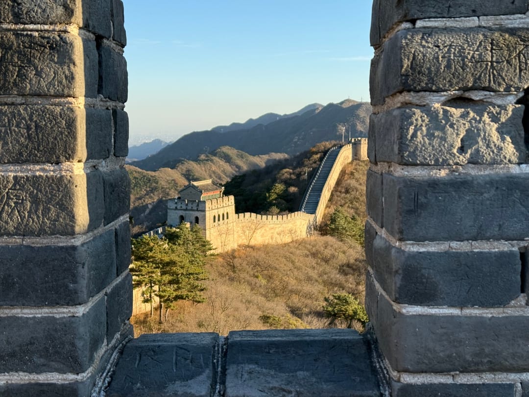 Blick durch eine Zinnenlücke der Mauer bei Badaling auf einen Wachturm und einen steilen Mauerabschnitt im Nachmittagslicht