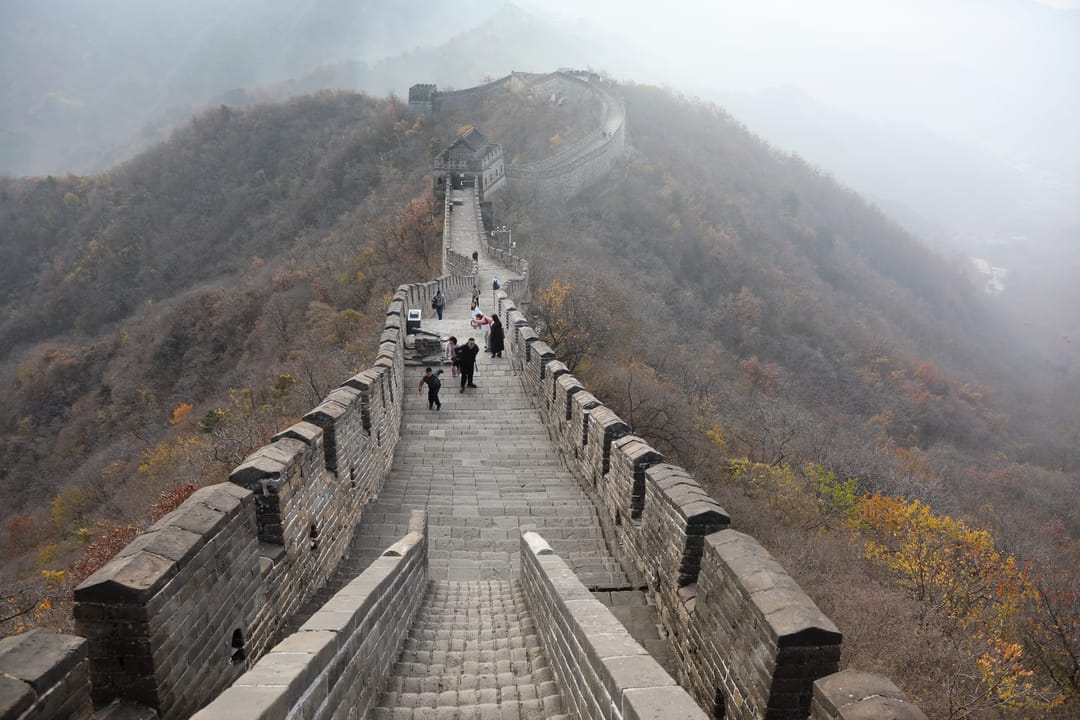 Il camminamento della Muraglia a Mutianyu, con merli su entrambi i lati, che segue una cresta autunnale verso una torre nella foschia