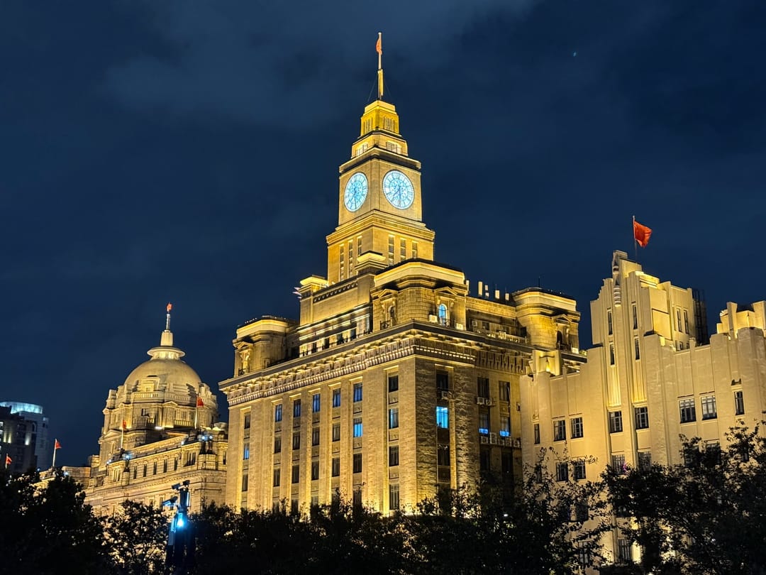 The Customs House clock tower floodlit in gold at night, with a domed bank building to its left, seen from below through trees