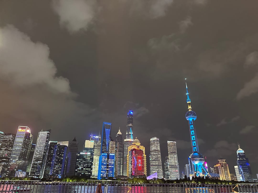 Lo skyline di Pudong illuminato di notte dal North Bund, con la Oriental Pearl Tower azzurra al centro e i grattacieli riflessi nel fiume