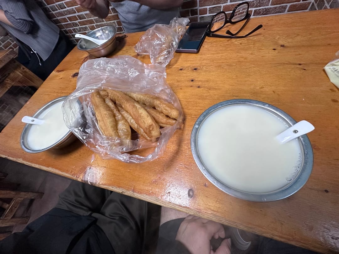 Youtiao fried dough sticks in a plastic bag between two metal bowls of soy milk on a wooden table