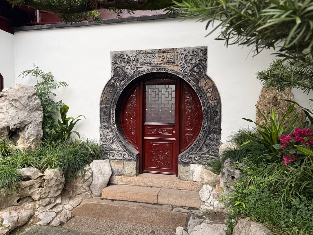 A round moon gate with a red wooden door set in a white wall in Yu Garden, framed by rocks and plants