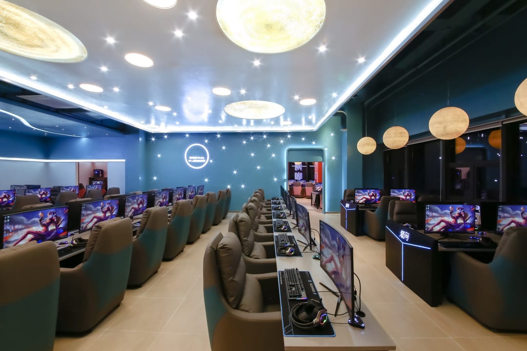 Filas de ordenadores para videojuegos y sillones reclinables bajo lámparas redondas en un cibercafé azul de Chengdu