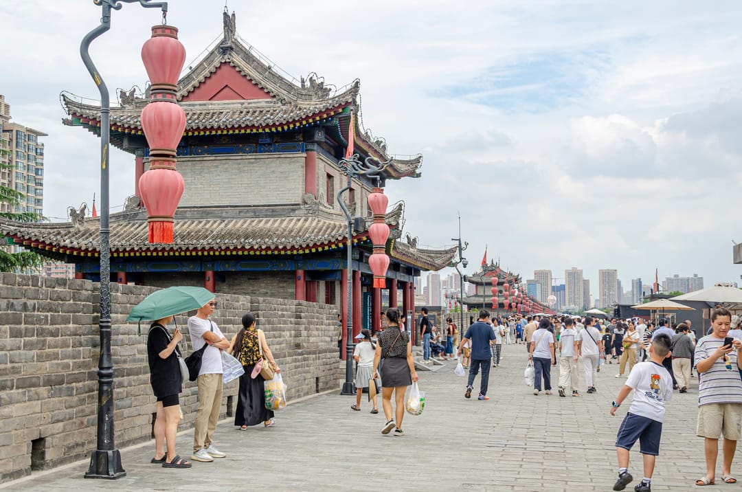 Visitantes caminando sobre la muralla de Xi’an junto a una torre de puerta con farolillos rojos y rascacielos detrás