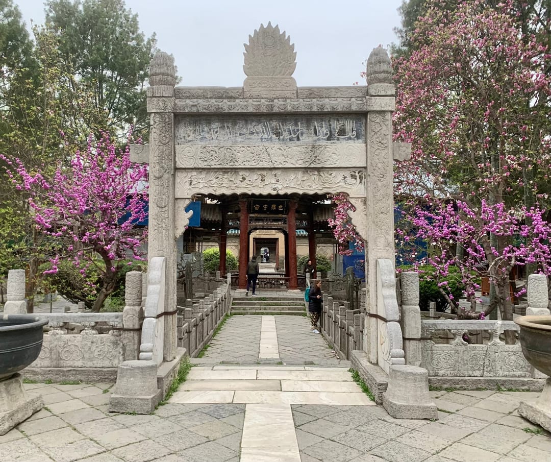 Una puerta de piedra tallada enmarcada por flores rosas en un patio de la Gran Mezquita de Xi’an, con una sala detrás
